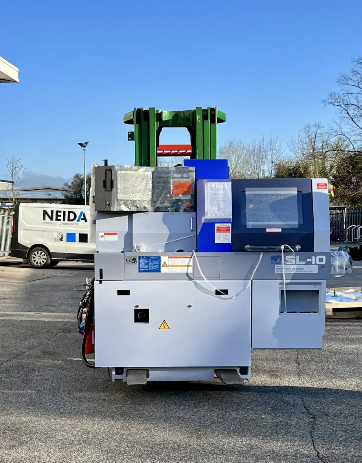A delivery scene showing the STAR SL10 sliding head machine being unloaded from a truck. The machine is securely fastened to a pallet and is being carefully lifted by a forklift operator. The delivery personnel are wearing protective gear and are working together to ensure a safe and successful delivery. The image captures the excitement and anticipation of receiving the advanced STAR SL10 sliding head machine, a valuable addition to the manufacturing facility.