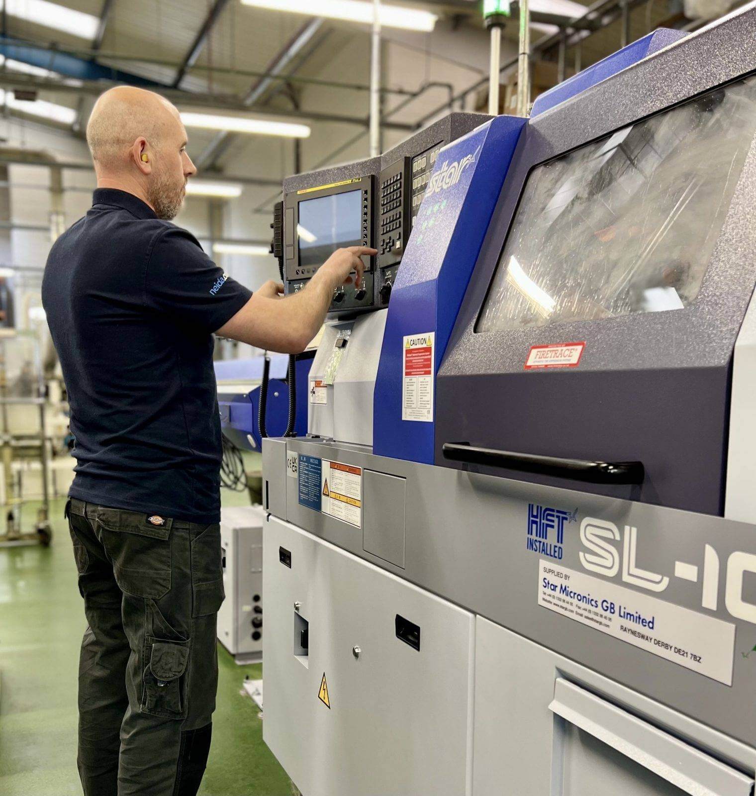 An employee wearing protective gear operating the new STAR SL10 sliding head machine. The employee is focused on the machine's controls, demonstrating technical expertise and precision. The machine is a modern and advanced piece of equipment used in manufacturing. The image showcases the employee's skill and dedication in utilizing the cutting-edge technology for efficient and precise production